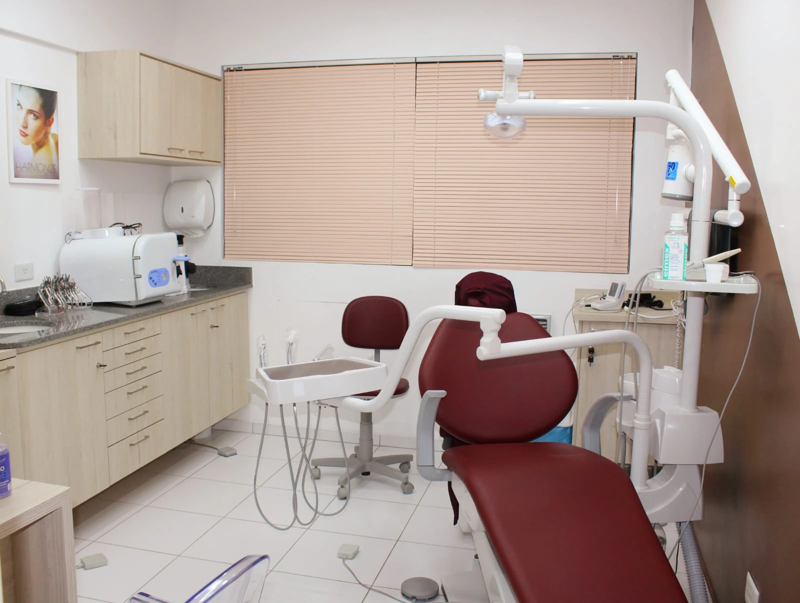 Sala de atendimento com cadeira odontológica bordô, armários de madeira clara e janela com persiana